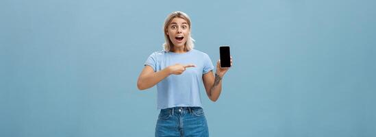 Studio shot of amazed and impressed charming european blonde in casual clothes gasping charmed at camera while showing smartphone screen pointing at device with finger over blue background photo