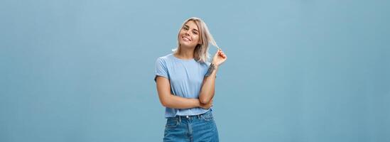 Studio shot of relaxed and chill popular attractive woman with blond hair and tattoos playing with strand tilting head and smiling as if listening boyfriend or flirting standing amused over blue wall photo