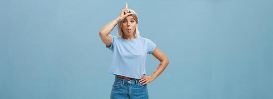 Studio shot of playful triumphing good-looking blonde female with tattoed arms showing letter L on forehead and sticking out tongue mocking over loser and celebrating own victory over blue wall photo