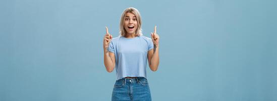 Waist-up shot of impressed enthusiastic creative blonde woman in trendy summer outfit smiling gasping being charmed and thrilled with awesome copy space pointing up over blue background photo