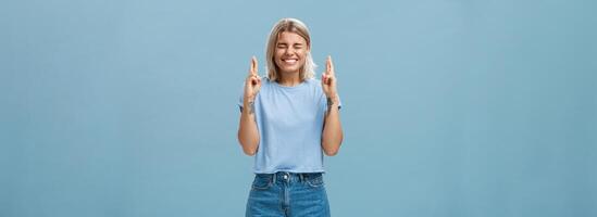 Optimistic faithful good-looking young woman with blond hair and tattoos smiling joyfully crossing fingers for good luck waiting for dream come true making wish while standing over blue background photo