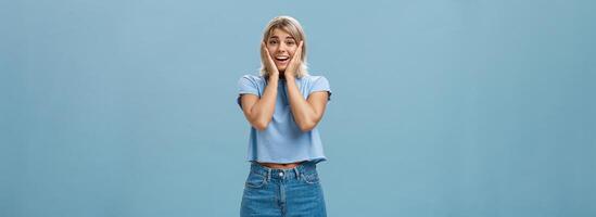 Oh mi entonces lindo. retrato de tocado oferta guapo atlético mujer con justa pelo participación palmas en las mejillas sonriente siendo impresionado y satisfecho en pie en mezclilla pantalones cortos terminado azul antecedentes foto