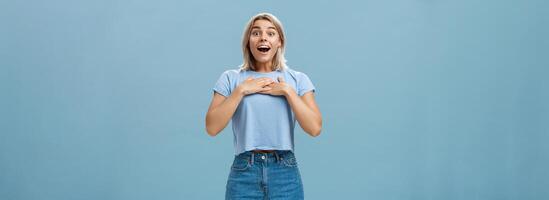 Portrait of amazed and charmed attractive blond female student with tanned skin in denim shorts and summer t-shirt holding palms on breast gasping and smiling joyfully being grateful and pleased photo