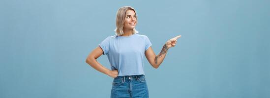 Studio shot of intrigued charming young european female with tanned skin and blond hair holding hand on waist in confident pose looking and pointing left with interest and curiosity photo