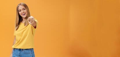 Woman directing at camera with index finger and smiling friendly as if talking to us standing amused and happy with confident carefree expression pulling arm towards to point over orange background photo