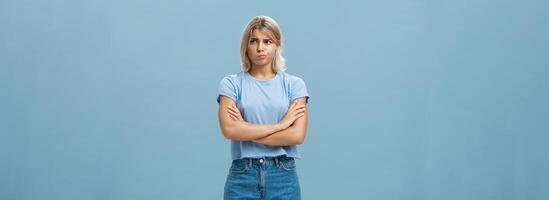 Uncertain troubled and perplexed attractive blond woman with tanned skin holding hands crossed on chest pouting and frowning looking right with worried unsure and doubtful look over blue wall photo