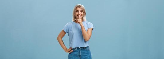 Studio shot of dreamy optimistic good-looking fair-haired female in denim shorts saying shh showing shush gesture with index finger over mouth smiling hiding secret over blue background photo