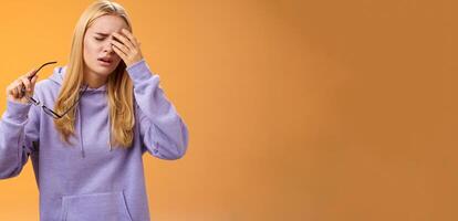 Tired dizzy cute blond girl taking off glasses touching forehead feel sick unwell suffering headache painful migraine standing bothered uncomfortable orange background in hoodie photo