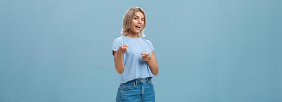 Hey I got offer for you. Portrait of attractive self-assured and playful stylish blond female with tanned skin in denim shorts and summer t-shirt winking and pointing at camera over blue background photo