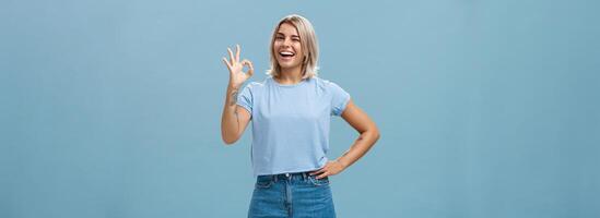 Confident and smart girl got everything under control. Portrait of optimistic good-looking girl with fair hair and tanned skin winking showing okay or perfect gesture standing over blue wall photo