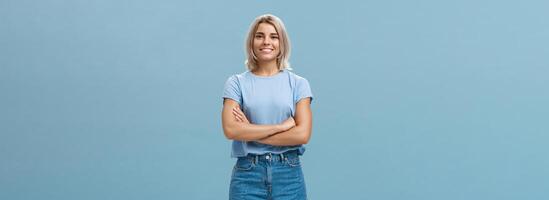 Ambitious good-looking young tanned female with blond hair standing in confident pose with hands crossed on chest smiling broadly standing in denim shorts over blue background photo
