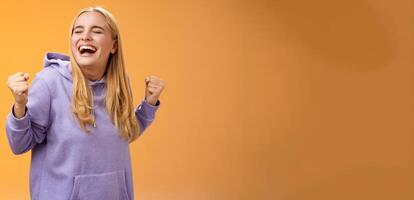 Cute happy triumphing blond european girl in hoodie yelling yes accomplish dream lifetime clench fists happily dancing celebrating victory success achieve goal, standing orange background photo