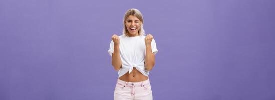 Rejoicing satisfied and pleased happy young ambitious girl with blonde hair in trendy white cropped top and shorts saying yes winking and smiling clenching fists in success or victory sign photo