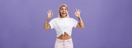 Girl being agree with friends deciding were enjoy holidays. Portrait of charming satisfied and happy young blonde female with tattoos and pierced belly showing okay gesture and smiling with delight photo