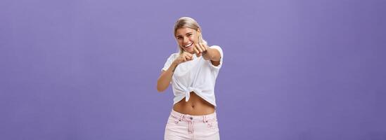 Ready fight back. Energized happy and awesome blonde female with tanned skin and fit body pulling fist in attack gesture towards camera smiling joyful acting like fighting or boxing over purple wall photo