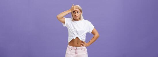 Woman got in serious bad sitation feeling concerned and nervous starting panic holding hand on forehead pursing lips staring down with shocked, perplexed expression being in trouble over purple wall photo