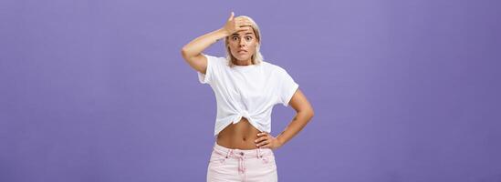 Damn I am in trouble. Worried troubled good-looking stylish female employee with tanned skin and fit body holding hand on forehead popping eyes from anxiety and making nervous face over purple wall photo