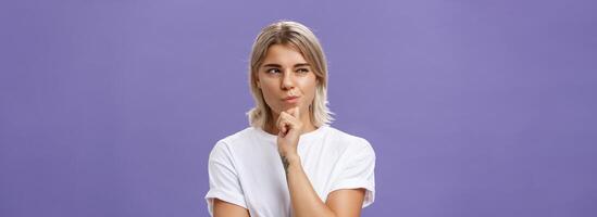 Waist-up shot of smart creative young ambitious woman with blond haircut and tanned skin squinting and smirking gazing left while holding hand on chin thinking making up plan or decision photo