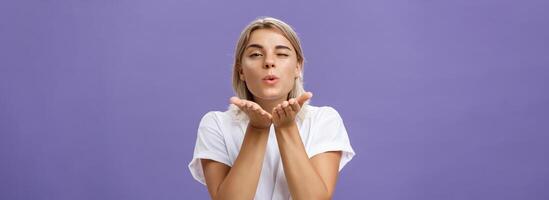 Blowing sweet kiss to all fans. Flirty tender and cute stylish european female with blond hair and tanned skin holding palms near mouth folding lips while sending mwah at camera over purple wall photo