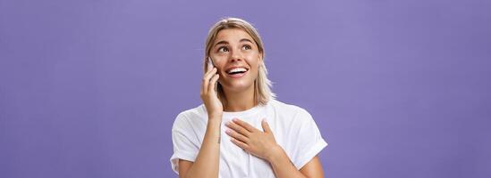 Waist-up shot of thankful charming blonde female student with tanned skin and blond hair holding hand on breast receiving delightful warm call from loving friend holding smartphone near ear gazing up photo