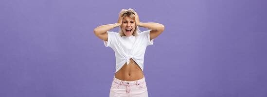 Portrait of panicking concerned and depressed young european woman with blond hair tattoos and athletic body yelling out loud holding hands on head being concerned and shook over purple background photo