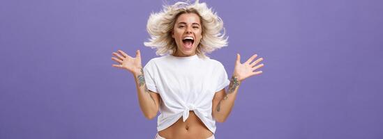 Studio shot of happy carefree and pleased joyful woman with blonde hair spreading palms aside in delighted pose smiling broadly and gazing entertained at camera jumping while having fun photo