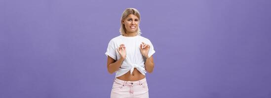 Ew take disgusting thing away from me. Stylish feminine good-looking blond woman in cropped top with tanned skin and pierced belly looking at creepy think with dislike and aversion over purple wall photo