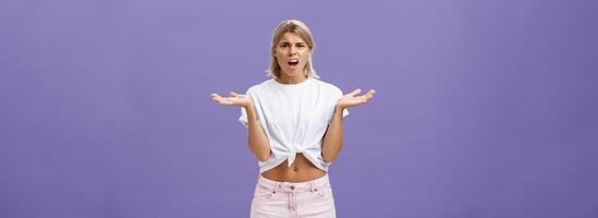Perplexed and disappointment good-looking blonde female student in white t-shirt and pink shorts frowning shrugging with spread hands near shoulders saying what the hell over purple wall photo