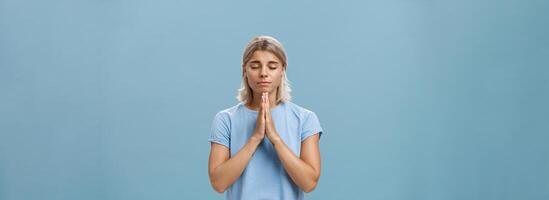 Good-looking kind and faithful european girl with blond hair smiling cute and tender holding hands in pray while making wish hopefully believing god hearing her prayers with closed eyes over blue wall photo