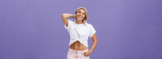 Indoor shot of carefree relaxed and joyful attractive athletic blonde woman in trendy summer outfit touching back of neck and smiling joyfully as if being in awkward situation over purple wall photo