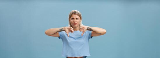 No way mate dislike. Portrait of dissatisfied bossy female tattoo artist with tattoos on arms frowning from displeasure showing thumbs down in disapproval standing over blue background photo