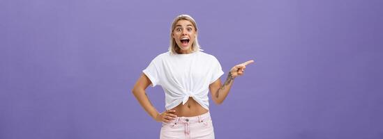 Thrilled and excited positive attractive woman with fair hair tanned skin and tattooed arms smiling joyfully with opened mouth staring impressed and delighted at camera, pointing left over purple wall photo