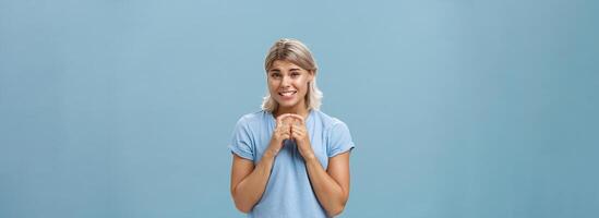 Silly cute and worried blonde female friend with short haircut steepling index fingers and smiling with guilty sorry smile frowning while waiting for answer begging for help or favor over blue wall photo