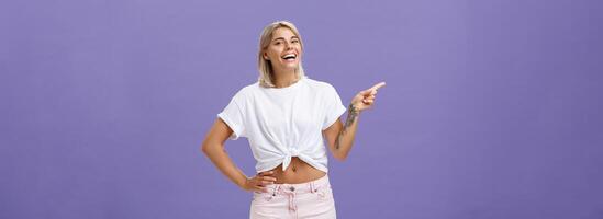 Check out cool place. Satisfied carefree stylish and attractive adult woman with perfect body tanned skin, tattoos laughing and smiling joyfully holding hand on hip and pointing left over purple wall photo