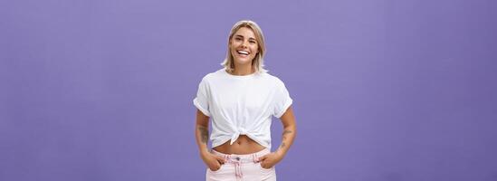 Joyful attractive tanned girlfriend with blond hair and tattoos on arms with cropped top chilling by pool with friends talking with amused happy expression smiling at camera holding hands in pockets photo