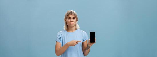 Unhappy sad attractive silly girl with fair hair tattoos and tanned skin frowning with regret and disappointment poonting at smartphone screen as if being dissatisfied with new girlfriend of ex-lover photo