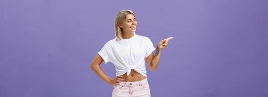 Hey nice job. Pleased carefree good-looking and confident female shop manager in stylish outfit holding hand on waist pointing and looking left with satisfied and delighted look over purple wall photo