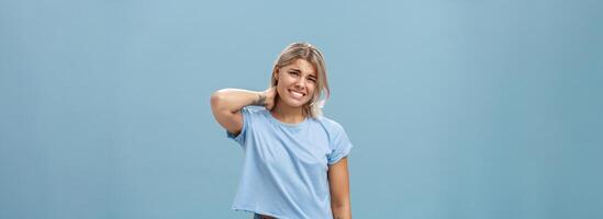 Girl unwilling to go with friend feeling awkward and unsure how say no frowning and clenching teeth making apologizing face rubbing neck behind saying sorry while rejecting offer over blue background photo