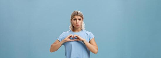 Heart being broken. Sad and gloomy heartbroken girl with blond hair tattoos on arms and tanned skin pursing lips whining and complaining making love sign over breast standing unhappy near blue wall photo