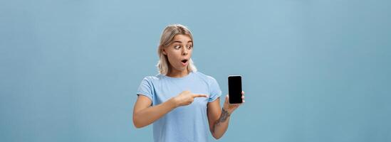 niña siendo asombrado con frio nuevo teléfono inteligente recibido en cumpleaños. sorprendido Sin palabras contento mujer con justa pelo y tatuaje goteante mandíbula participación teléfono inteligente señalando y curioso a artilugio pantalla foto