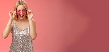 Sassy carefree daring young blond girl winking cheeky smiling assertive making first move approach handsome man nightclub asking dance standing red background touching sunglasses wear dress photo