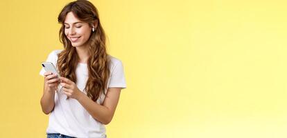 Happy caring tender feminine girlfriend curly long hair hold smartphone picking song listen way home smiling broadly look mobile phone scroll music platform wear wireless earbud yellow background photo