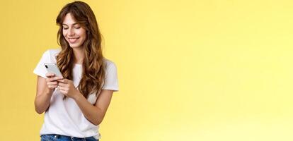 Cheerful lovely girlfriend texting friend pleased cute smile tap smartphone screen smiling broadly look mobile phone display tenderly writing post contemplate touching photo yellow background