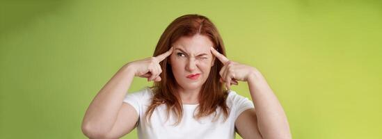 Difficult decision. Perplexed hesitant redhead middle-aged 50s woman trying solve puzzle look puzzled troubled smirk close eyes peek right touch temples thoughtful thinking intense green background photo