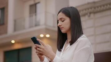 vrouw knikken terwijl typen een bericht Aan smartphone in voorkant van kantoor gebouw. video