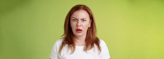 Confused shocked gasping middle-aged redhead woman cringe frustrated puzzled open mouth speechless freak out strange shocking scene stand green background perplexed disappointed photo