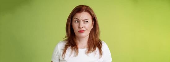 Perplexed doubtful unsure redhead wife smirking unsatisfied frowning look aside displeased disappointed hesitating pondering strange advice thoughtfully stare away stand green background photo