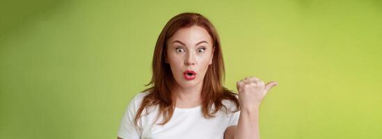 Close-up surprised amused redhead middle-aged housewife folding lips say wow impressed stare camera questioned ambushed pointing left thumb astonished wondering if promo true green background photo