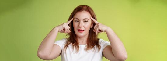 Smart choice. Intrigued thoughtful good-looking assertive redhead creative middle-aged woman touch temples squinting pondering info thinking use mental power read minds stand green background photo