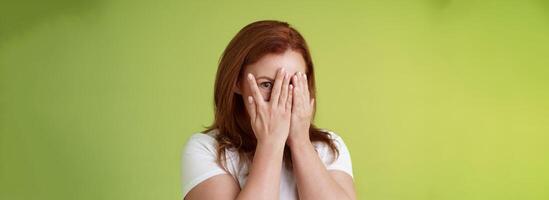 Not peeking. Playful charismatic middle-aged ginger redhead woman close face palms look through fingers one eye stare camera intrigued waiting surprise gift stand green background photo